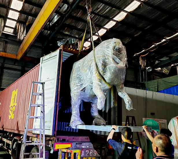 Large wrapped dinosaur being moved by crane beside a shipping container